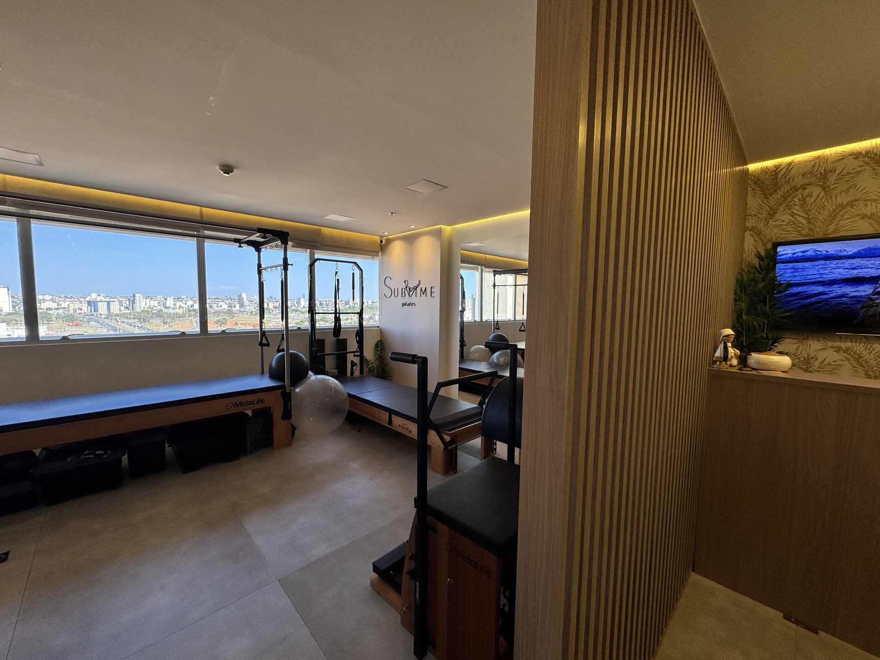 Vista ampla do estúdio Sublime Pilates: reformers alinhados junto à janela com vista da cidade, logo na parede e painel ripado