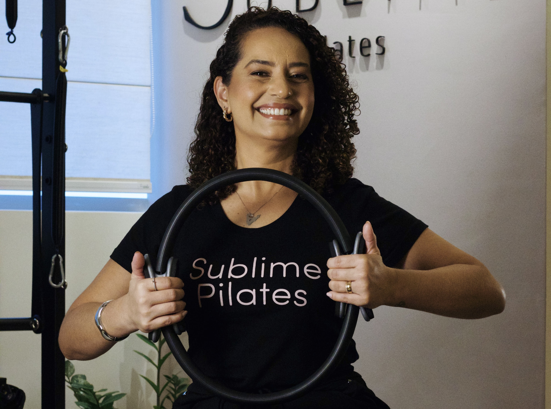 Paula Araújo, fisioterapeuta e fundadora do Sublime Pilates, sentada em um reformer com o logo do estúdio ao fundo