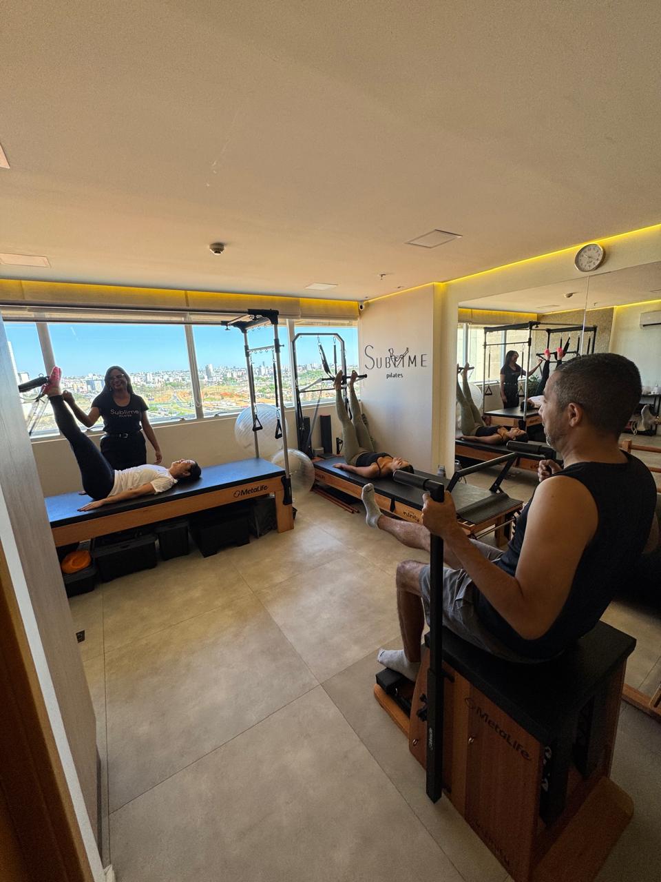 Aula de Pilates com turma reduzida no Sublime: instrutora acompanha alunos em reformers, com o logo do estúdio e a vista da cidade ao fundo